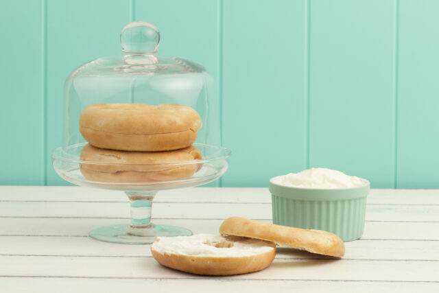 A bagel spread with cheese cream on a white wooden table. A turquoise bowl with cheese cream and two bagels in a cake stand with a cloche.